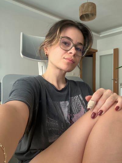 The image displays a young adult female seated indoors, likely in a residence or office in Karşıyaka, Turkey. She faces the viewer directly, exhibiting a neutral expression with slightly puckered lips. Her medium-brown hair is casually styled, and she wears olive-green framed glasses and small gold hoop earrings. A gold chain necklace is visible around her neck. She is dressed in a dark grey t-shirt featuring a faded graphic print depicting architectural structures, resembling the Colosseum, alongside a stylized purple figure. Her right hand rests on her right knee, revealing dark red nail polish on her visible fingers. The index finger of her right hand is wrapped in a light-colored bandage. Her exposed legs suggest she is wearing shorts or a skirt. Behind her, a modern light grey chair with a distinctive backrest is partially visible. The background consists of a light-colored wall. To her right, a light brown wooden doorframe leads to a white door with a frosted glass panel, complete with a metal handle. A cylindrical light fixture, appearing to be constructed from woven material, hangs from the ceiling. A fragment of a green plant leaf is visible on the extreme right edge of the frame.