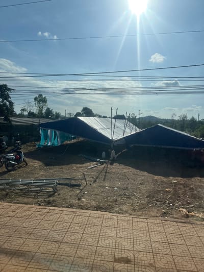 The image depicts an outdoor scene in Xuân Long, Vietnam, under a bright blue sky with scattered clouds and intense sun glare. Numerous black utility lines traverse the upper frame. In the foreground, a patterned light-brown concrete sidewalk is visible. Beyond it lies an unpaved, brown dirt lot containing two large, temporary canopy structures, featuring dark blue tarp roofs and white translucent sections, appearing to be in a state of assembly or disassembly. Light blue and white fabric hangs from the side of one canopy. Several metal support poles are evident, and multiple sections of metal scaffolding are scattered on the ground in the lower-left. At least two individuals, wearing light-colored clothing, are present near the canopies, engaged in activity related to the structures. A white scooter with red accents is parked on the left side of the dirt lot, near a dark fence. Green trees and distant hills are visible in the background.