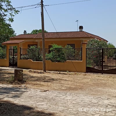 This is a medium-shot, eye-level photograph of a single-story house in Grumo Appula, Italy, on a clear, sunny day. The house has a yellow stucco exterior and a terracotta tile roof. A black metal fence encloses the property, with a black metal gate on the left side leading to the entrance. To the right, there is another black metal gate. The yard is dry and dusty, with patches of dry grass and a paved area in the foreground made of irregular, flat stones. A wooden pole with electrical wires runs vertically on the left side of the frame, and a second, thinner pole is visible on the right. There is a small tree stump in the foreground to the left. The image contains a timestamp in the lower right corner: "16.07.2026 14:12".