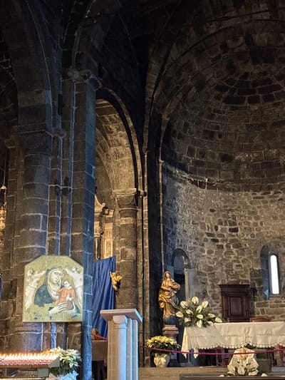 The image displays the interior of the Church of Santa Margherita d'Antiochia in Vernazza, Italy. The environment is characterized by large, dark, rough-hewn stone blocks forming massive structural elements, including square and rectangular pillars, pointed arches, and a vaulted, domed ceiling. The lighting is dim, suggesting a solemn atmosphere, with light sources localized to specific areas and a narrow, arched window visible on the right wall.
In the left foreground, a multi-tiered metal stand holds numerous lit votive candles. Adjacent to this display are potted white poinsettia plants. Above, affixed to a stone pillar, is an octagonal painting depicting the Virgin Mary in a dark blue robe holding the infant Jesus. Faint angelic figures are visible in the background of the artwork.
Towards the center, a light-colored fluted stone pedestal stands in the mid-ground. Behind this pedestal, a section of dark blue fabric is draped against a stone wall or niche. To the right of the fabric, a small golden figure, appearing to be a cherub or angel, is mounted on the wall.
Further to the right, a golden statue of a robed female figure, positioned on an ornate red and gold pedestal, stands prominent. In front of this statue, an altar table is covered with a white tablecloth featuring lace trim, embroidered patterns, and a red braided cord along its edge. On the altar, floral arrangements of white peace lilies or similar flowers are present, alongside another potted white poinsettia. To the right of the altar, a dark wooden cabinet or piece of liturgical furniture is situated against the stone wall beneath the arched window.