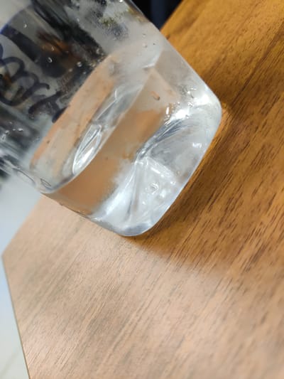 The image is a close-up, angled shot of a clear plastic bottle filled with a clear liquid, resting on a wooden surface. The bottle's bottom corner is visible, showing condensation on the outside. The liquid inside has some slight opaqueness, possibly due to dissolved substances or air bubbles. The wooden surface has a visible grain pattern and appears to be a tabletop. The background is out of focus, showing some dark and light shapes that are not clearly identifiable. There are no people or discernible actions taking place in the image. The image does not provide any information to determine its location in Neftekachka, Russia.