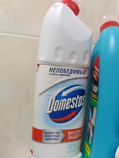 The image shows two cleaning product bottles, likely toilet bowl cleaners, standing next to each other against a beige tiled wall. The bottle on the left is white with a red cap and a label that prominently features the brand name "Domestos" in large blue and white letters. Text on this label includes "НЕПОБЕДИМЫЙ" (Invincible) and "В БОРЬБЕ С МИКРОБАМИ" (In the fight against microbes). Below the Domestos logo, there is text in Russian that translates to "KILLS ALL KNOWN MICROBES" and "MAXIMUM PROTECTION". The bottle on the right is bright blue with a red label. A portion of its label is visible, showing text that appears to be in Russian as well, and possibly the brand "Sanfor". The context does not suggest a specific location in Jitiqara, Kazakhstan, as there are no discernible landmarks or identifying features related to that city.