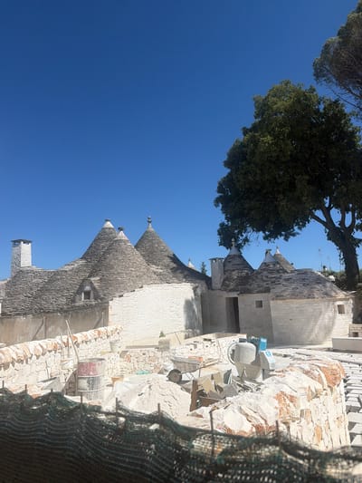The image depicts a construction site in Alberobello, Italy, showcasing traditional Trulli houses under renovation or construction. The Trulli, characterized by their conical roofs made of stone slabs and whitewashed walls, are a prominent feature of the landscape. In the foreground, construction materials such as sand, stones, and a cement mixer are visible, indicating ongoing work. A red and black barrel and a wheelbarrow are also present. A stone wall, partially constructed, encloses the area. Beyond the construction zone, the unique architecture of multiple Trulli houses stands against a clear blue sky. A large, leafy tree is situated to the right of the Trulli, casting a shadow over some of the structures. The overall scene suggests a restoration or development project of Trulli buildings in Alberobello.