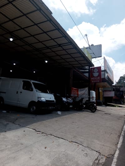 The image shows a street scene with a Mixue store and various vehicles parked under a corrugated metal awning. A white van is parked closest to the camera, followed by a black car and two motorcycles. A Mixue sign is visible in front of the store, and recruitment banners for riders and drivers can be seen. The ground is concrete with cracks and debris. The sky is partly cloudy. The license plate number of the white van is 6608 PO. The location is inferred to be in a city, likely Kioumaknao 2, Laos.