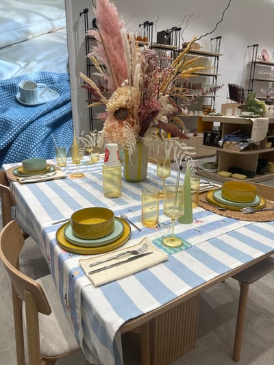 The image depicts a table setting inside a store in Родниковское сельское поселение, Ukraine. The table is covered with a blue and white striped tablecloth, and place settings for two people are visible. Each setting includes a yellow bowl, a green plate, a yellow plate, silverware, and a napkin. Woven placemats sit beneath the plates. Two yellow-tinted glasses are set on either side of a yellow-striped vase, which holds a large, dried flower arrangement. In the background, shelving units display various housewares and decorative items. A couch or bed is partially visible on the left side of the frame. Chairs are situated on opposite sides of the table.