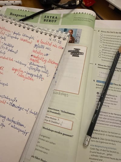 The image shows a close-up view of a desk with several items. In the foreground, a black pencil with a silver eraser is resting on an open book. The book's pages contain German text, with some sections highlighted and handwritten notes in Armenian script. To the left of the book, a spiral-bound notebook is open, also filled with handwritten notes in Armenian script. The notebook is positioned on top of a document with the heading "Thema" and "Besprech". To the right of the pencil, another open book displays German text and a multiple-choice question with checkboxes. The background is out of focus but suggests a cluttered workspace. The scene is indoors, and there are no visible indications of the city of Sharjah, United Arab Emirates.