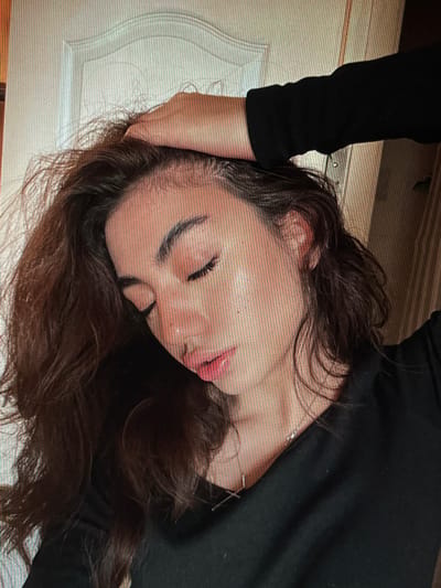 A young woman with long, dark, wavy hair is shown in a close-up, chest-up portrait. She is wearing a black long-sleeved shirt and a delicate silver necklace. Her left hand is resting on her head, and her eyes are closed, with her head tilted downwards. Her lips are slightly parted and appear to be glossed. The background is indistinct, but suggests an indoor setting with a door visible on the left. There is no discernible information within the image that would associate it with Chotěšov, Czechia.