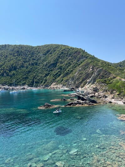 The image depicts a scenic coastal view with clear turquoise water, rocky outcrops, and a verdant, tree-covered hillside under a bright blue sky. Several sailboats and a small motorboat are anchored in the calm bay. The coastline is characterized by rugged terrain with visible rock formations and sparse vegetation on the slopes. In the foreground, the shallow, transparent water reveals the rocky seabed. The overall impression is one of a serene and picturesque Mediterranean landscape, likely a secluded cove or bay. The mention of Loutraki, Greece, suggests this location might be a popular tourist destination or a natural attraction in that region.