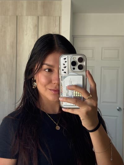 A young woman is captured in a mirror selfie, holding a smartphone with a clear case that contains several cards and black star stickers. Her head is tilted, and she gazes directly into the mirror with a calm expression, showing a subtle smile. She has long, dark hair, brown eyes, and is wearing a black top.

Notable accessories include a gold-toned ring on her right index finger, a black elastic-like bracelet, and a delicate gold-toned chain bracelet with a small charm on her right wrist. A gold-toned necklace with a small, light pink or lavender hexagonal pendant is visible around her neck, and she wears a decorative gold-toned earring on her right ear.

The setting appears to be indoors, potentially in a bedroom or hallway, indicated by a white panel door with a doorknob in the background. To the left, a wall features light-colored horizontal wood paneling. The lighting is soft and even, suggesting an indoor environment during daytime or with good artificial illumination.

Visible text from the cards held within the phone case includes reversed and fragmented characters, some resembling "X", "2 Бперія", "Glastoire Mوروالى", "800 33 11 100", "Qush hutisilor", "ม.www", "261 eb bsbinevir", "ДА П62 3ЛЬМ ЕπίπεJε", "+25 (333) 530 30 00", and "ИЕ". These fragments likely pertain to personal or commercial information on the cards. The activity takes place in San Francisco Ocotlán (Ocotlán), Mexico.