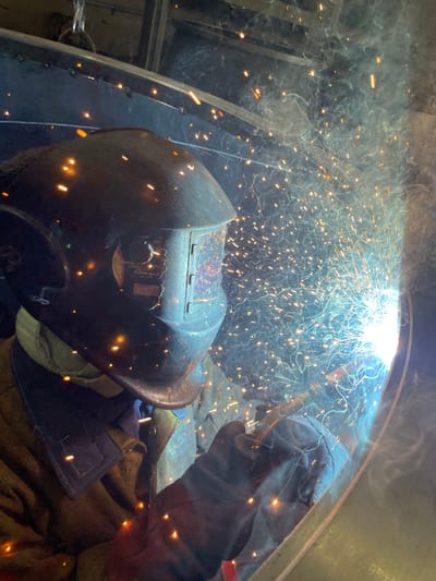 A person in protective welding gear, including a dark helmet and heavy gloves, is performing a welding operation. Sparks and smoke are filling the air, emanating from the bright point of the welding arc. The welding is taking place inside what appears to be a metal structure. The specific location of the city of Tomba, Montenegro, is unknown in the scene.