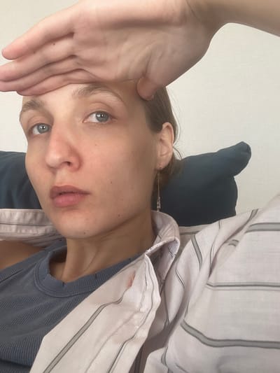 A woman is seen indoors. She is touching her forehead with her hand. She appears to be resting against a pillow, wearing a blue tank top and a striped shirt. The environment is plain and simple, the background is white.
There is no information given in the image to confirm that it was taken in the city of Sepych, Russia.