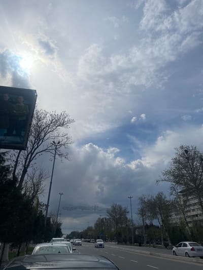 The image depicts a view in Navoiy City, Uzbekistan, showcasing a sky with scattered clouds and patches of blue. Buildings and trees line a road with parked cars. An advertisement or display is visible in the foreground, along with streetlights and overhead power lines stretching into the distance.