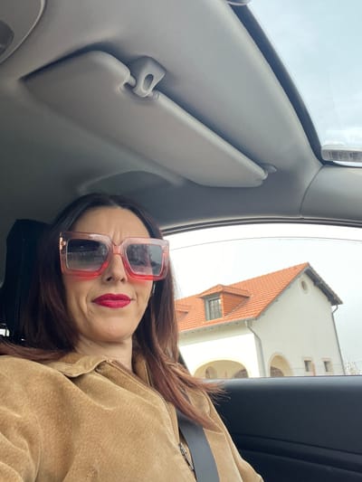 The image shows an adult female positioned in the driver's seat of a vehicle, looking towards the viewer. She has dark brown hair and is wearing oversized, square-framed sunglasses with a clear pinkish-red gradient frame and dark lenses. Her lips are coated with bright red lipstick, and she displays a neutral to slightly contemplative expression with a furrowed brow. She wears a tan or light brown textured jacket with a visible zipper closure down the front, and a dark seatbelt is diagonally across her chest. The vehicle's interior includes a light gray headliner, with a sun visor in the raised position on the passenger side. Through the passenger window, a distinctive building is visible under an overcast sky. This building is the former railway station (Gare de Roodt-sur-Syre) in Roodt-sur-Syre, Luxembourg. It features a light cream-colored facade, a prominent steeply pitched gable roof covered with orange terra cotta tiles, and a dormer window with dark framing on the visible gable end. The ground level of the building presents a series of three distinct arched openings, and a circular decorative element is visible on the upper facade near the roofline on the right side.