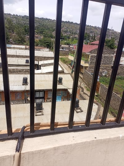 The image presents an elevated, downward-angled view of a residential area in Kiamaina ward, Kenya, framed in the foreground by black vertical metal bars and a cream-colored concrete balustrade. A light-colored cable, taped with black material, is visible on the balustrade.

The midground features numerous single-story buildings with corrugated metal roofs. Multiple black cylindrical water tanks are observed on these roofs, some positioned on wooden platforms. Building exteriors appear to be constructed from rough stone or concrete blocks, with visible windows, some featuring metal grates, and doors, including a black metal gate and some blue-painted doors. Narrow, unpaved dirt paths weave between these structures. Further to the right, a two-story building with yellow walls, a red tiled roof, and a green-railed balcony is partially visible.

The background shows a densely built-up, hilly landscape covered with trees and additional houses extending upwards. A communication tower is visible on a distant hill to the right. The sky is overcast. No individuals are present in the frame.
