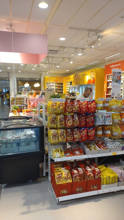 The image shows the interior of a supermarket aisle in Pylaia Municipal Unit, Greece. In the foreground, a refrigerated display case containing food items is visible on the left. To the right, shelves are stocked with various packaged snacks, including bags of "Giffles" cookies and "Dunle" cookies, as well as boxes of yellow-packaged snacks. A man with glasses is standing behind the shelves, appearing to be working or shopping. In the background, there are more shelves with products, a yellow wall with graphics and text, and lighting fixtures on the ceiling. The overall environment suggests a retail setting focused on food and snacks.