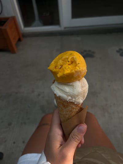 A person holds an ice cream cone with two scoops of ice cream in their hand. The cone is wrapped in brown paper. One scoop is yellow and the other is white. The person's fingers are visible holding the cone. The background shows a gray wall with paw prints and a window with a brown wooden stool. The setting is likely outdoors in La Esperanza, Honduras.