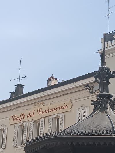 The image shows a building facade in Casalmaggiore, Italy, with the signage "Caffè del Commercio" prominently displayed. The building features several windows with white shutters. In the foreground, a decorative metal structure, possibly part of a fountain or a kiosk, is visible. The sky above is clear and pale blue. The architectural style suggests a historic European setting. There are no people visible in the image. The scene is captured from a low angle, looking upwards towards the building and the sky.