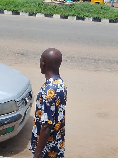 A medium shot captures a bald Black man from the chest up, facing away from the camera and looking to his right. He is wearing a dark blue short-sleeved shirt with a floral pattern featuring yellow and white roses. He is standing next to a silver car, with the front left side of the car visible to the left of the man. The car appears to have some wear and tear, with scuff marks on the bumper. The ground is covered in sand and dirt. In the background, a paved road is visible, bordered by a curb with alternating black and white stripes, beyond which is a patch of green grass. Further in the background, a yellow motorized rickshaw and a few people are faintly visible. The scene appears to be an outdoor urban or semi-urban environment.