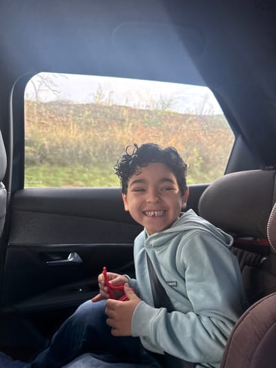 The image captures a male child, estimated between 6 and 9 years old, seated in the rear passenger compartment of a vehicle in Pourrières, France. The child has dark, curly hair and is wearing a light blue hooded sweatshirt with a 'Levi's' brand patch on the left chest, over blue denim trousers. He is secured by a black shoulder seatbelt positioned diagonally across his torso. The child is smiling widely, facing the camera, and several lower front teeth are visibly absent. Both hands are engaged with a red, flexible, pipe cleaner-like object or toy, shaped into an abstract, multi-looped configuration. The vehicle's interior includes a dark door panel with a silver-toned door handle visible on the left, and a segment of a brown-upholstered car seat or booster seat on the right. Through the passenger window, a natural landscape is visible under overcast conditions, featuring a sloped area covered in dry, light brown grasses and sparse, bare-branched vegetation, indicative of a dormant season.