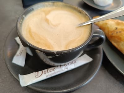 The image displays a close-up view of a cup of coffee with latte art, resting on a dark saucer. A silver spoon is partially submerged in the frothy coffee. To the left of the cup, two small, white packets are visible on the saucer, one of which appears to be a sugar packet with text. In the background, a pastry with sesame seeds is out of focus, and another small white object, possibly a pastry wrapper, is visible on a separate plate. The setting appears to be a cafe or restaurant, suggested by the presentation of the coffee and pastry. The lighting suggests it could be daytime. The text on the visible packet reads "Dolce Parma" and "aria patisserie".