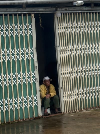 An elderly man wearing a white cap, yellow jacket, and green pants sits on a low platform inside a doorway in Tu Póh, Vietnam. The doorway is partially obscured by a collapsible metal gate with green and white vertical panels. The man is facing right, with his hands resting on his knees. The ground in front of the doorway is wet, suggesting recent rain or puddles. The environment appears to be an outdoor setting, possibly a shop or residential entrance, characterized by a simple, utilitarian structure.