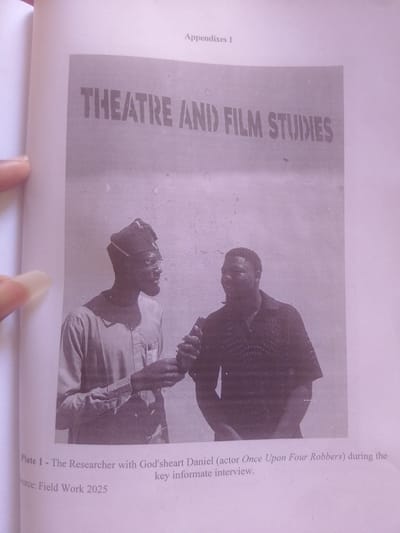 The image is a black and white photograph taken from a book, likely a thesis or academic publication. The photograph itself is a black and white plate showing two men in what appears to be an informal setting. The text above the photograph indicates it is part of "Appendixes 1" and the title displayed on a textured background reads "THEATRE AND FILM STUDIES."

The two men are positioned centrally in the frame. The man on the left is wearing a traditional cap and a light-colored, buttoned shirt. He has glasses and a beard and is looking towards the right. His left hand is raised, holding something small, possibly a recording device or microphone. The man on the right is wearing a dark, patterned shirt. He is facing the left and appears to be speaking, with his mouth slightly open. He has a darker complexion and is more heavily built than the man on the left. The background is a plain, textured wall.

Below the photograph, there is a caption that reads: "Plate 1 - The Researcher with God'sheart Daniel (actor Once Upon Four Robbers) during the key informate interview. Source: Field Work 2025." This caption provides crucial context, identifying the individuals as a researcher and God'sheart Daniel, an actor from "Once Upon Four Robbers," engaged in an interview. The "Field Work 2025" suggests the photograph was taken as part of research conducted in that year. The location context provided is Magajin Gari, Nigeria, which suggests the interview and field work took place in that region. The overall tone of the image is documentary and academic.