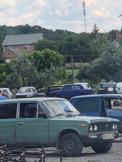 The image depicts a street scene in Pidhaitsi, Ukraine, on a cloudy day. In the foreground, a green car is parked on a gravel surface, with a portion of its front visible. The car has a license plate that reads "BA 5587 AP". Behind the green car, a dark blue car is parked. Further in the background, several other vehicles are visible, including a light-colored car and a dark-colored sedan. Beyond the cars, there is a dense line of trees, and behind the trees, a house with a red brick facade and a grey roof can be seen. A tall telecommunications tower stands in the background, partially obscured by trees. The overall impression is of a quiet, residential area with a collection of parked vehicles.