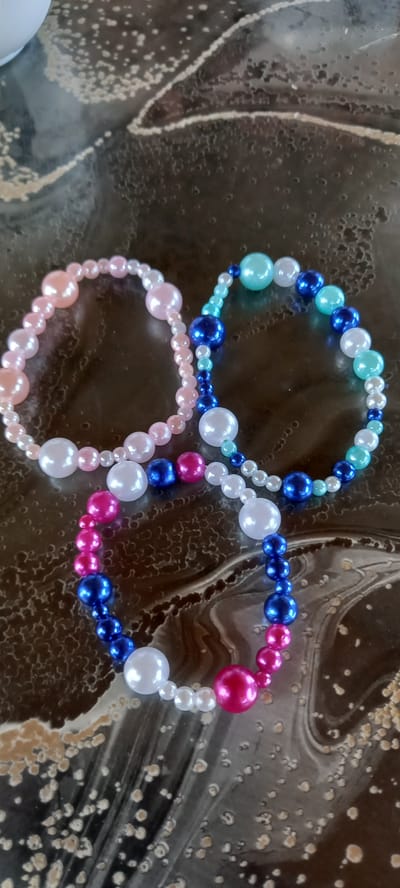 Three beaded bracelets are arranged on a dark, textured surface. The bracelet on the left is composed of light pink and pearl white beads. The bracelet in the middle is a mix of aqua, dark blue, and pearl white beads. The bracelet on the right features dark blue, bright pink, and pearl white beads. The surface appears to be a tabletop with a marbled or patterned finish. The lighting suggests an indoor setting. The image does not contain any information about the city of Iskra, Bulgaria.
