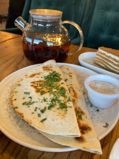 The image shows a table setting with food and drinks on a wooden table. In the foreground, on a white plate with a textured rim, are two folded crepes topped with chopped green herbs, and a small white ramekin filled with a creamy white sauce. To the right of the crepes, on a separate oval white dish, is a slice of layered cake. In the background, a glass teapot filled with a dark red liquid, presumably tea, sits on the table. The teapot has a wooden lid and handle. Behind the teapot, a dark green upholstered chair is visible. The scene is indoors, likely in a cafe or restaurant. The image captures a moment of a meal or snack. There are no people visible in the frame. The location is identified as Vysokaya Gora, Russia, suggesting this scene is taking place in a dining establishment in that city.