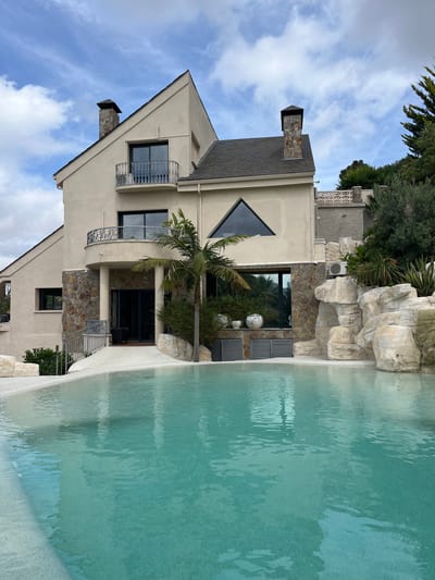 The image depicts a large, modern house with a swimming pool in the foreground, located in Can Güell, Spain. The house has a stucco exterior in a light beige color, with a dark grey tiled roof and several chimneys. The architecture features a mix of sharp angles and rounded elements, including balconies with ornate metal railings. The house is partially clad in stone on the lower levels. A palm tree stands in front of the house, near the entrance. The swimming pool has a light-colored, textured surround and is filled with clear turquoise water, reflecting the sky. Behind the house, there are rocky features that resemble a natural landscape, and lush green vegetation. The sky is partly cloudy with scattered white clouds against a blue backdrop. There are no people visible in the image.