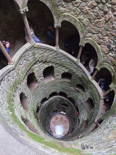 This image depicts the interior of the Initiation Well (Poço Iniciático) at Quinta da Regaleira, located in Sintra (Santa Maria e São Miguel, São Martinho e São Pedro de Penaferrim), Portugal.
The scene is an aerial view looking directly down into a deep, inverted stone tower featuring a wide, spiraling staircase that descends into the earth. The well's walls are constructed from rough-hewn, grey stone, heavily textured and covered in patches of green moss and lichen, particularly on the ledges and arches.
Numerous individuals are present within the structure at various levels of descent. In the upper left quadrant, a person in a blue shirt holds a black smartphone horizontally, aiming it towards the well's interior, consistent with taking a photograph. Adjacent to them, another individual in a light-colored top and blue skirt stands near an arched opening. Several other people, dressed in casual clothing including jackets and trousers, are distributed along the spiral staircase and within the arched passageways, either descending or ascending.
The spiral staircase features multiple arched openings and windows along its inner wall. At the deepest visible point of the well, a circular floor is discernible, adorned with a geometric pattern, possibly a compass rose, and partially covered by a shallow layer of water that reflects ambient light. The overall lighting is subdued, with shadows cast within the deep recesses and beneath the arches, indicating either natural light from above or artificial illumination not directly visible. The perspective emphasizes the structure's depth and winding nature.