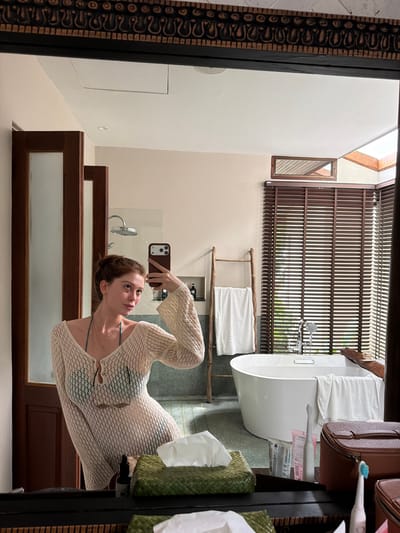 A woman with red hair pulled back in a bun takes a mirror selfie in a bathroom. She is wearing a light-colored, sheer, knitted cover-up over a dark-colored bikini top. She is holding a smartphone in her right hand, with the camera facing the mirror. In the background, a white bathtub is visible, with a wooden ladder leaning against it and a white towel draped over it. Blinds cover a window to the right of the bathtub. The bathroom appears to be in a resort or hotel, possibly located in Ao Hin Khok, Thailand, suggested by the presence of tropical plants visible through the blinds and the overall aesthetic of the room.