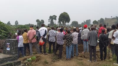 A large group of people are gathered outdoors in a rural, hilly area with trees. They seem to be standing around what might be a grave or burial site. The scene appears to be overcast or foggy. Some of the people are wearing casual clothing. There is a tombstone to the left with a picture attached. The crowd looks solemn.