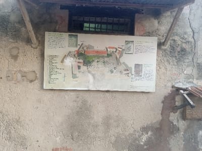 A weathered, light-colored plastered wall, exhibiting significant discoloration, cracks, and patches of exposed substrate, is depicted in the Makadara/Mji Wa Kale ward, Kenya. Centered on this wall is a large, rectangular informational sign, primarily white, featuring an illustrated map or aerial rendering of a historical settlement, identified by text including "Citta Vecchia Mombasa" (Old Town Mombasa). The sign also contains multiple columns of descriptive text and smaller inset images or diagrams. Above the sign, a rectangular window with dark, horizontal bars reveals a shadowed interior. An angled corrugated metal overhang projects above the window, supported by visible wooden struts, one extending from the upper left portion of the wall towards the top edge of the frame. To the lower right of the sign, a smaller, dark L-shaped bracket with a terracotta-colored cap is affixed to the wall. No individuals are present in the scene.