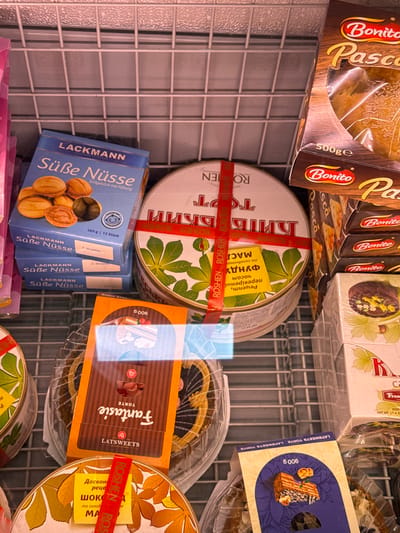 This is a close-up, slightly distorted, eye-level shot of various sweet food items displayed on a metal shelf, likely in a supermarket or bakery in Lloret de Mar, Spain. The primary subjects are packaged cakes and cookies, with a focus on baked goods with chocolate, nut, and fruit fillings.
In the foreground, a blue box labeled "LACKMANN Süße Nüsse" with an image of walnut-shaped cookies is prominent. To its right, a transparent plastic container holds a chocolate cake, partially visible, with an orange wrapper featuring the word "Fantasie" and "LATSWEETS TORTE." Another circular tin with a red ribbon and green leaf design, labeled "ROSHEN," is also visible, along with a yellow sticker with text in what appears to be Cyrillic script. In the background, several chocolate cookies with nut and fruit toppings are stacked in their packaging, some with the brand name "Bonito."
The scene is brightly lit, suggesting an indoor commercial setting. The metal shelving grid is a recurring element throughout the image, providing a sense of depth and context. The overall impression is one of abundance and variety in confectionery offerings. No people are visible, and the focus is solely on the products. The time of day and weather are not discernible from the image.
