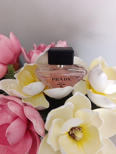 A perfume bottle is placed among artificial flowers. The bottle is clear glass with a pinkish liquid inside and a black cap. The brand name "PRADA MILANO" is visible on the bottle. The flowers are large and appear to be magnolias, with pink and yellow and white petals. The background is a plain, light gray wall. The image was taken indoors in a well-lit environment. There is no indication of the location in Toruń, Poland, nor are there any people or actions depicted.