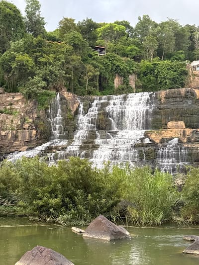 The image shows the Pongour Waterfall in Tân Phú, Vietnam. The waterfall cascades down a series of rocky tiers, with lush green vegetation surrounding it. The water is a murky green color, and there are several large rocks in the foreground. The sky is overcast, and the overall scene is one of natural beauty and tranquility.