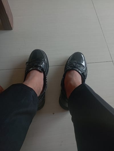 In the city of Batulawang, Indonesia, an overhead perspective captures the lower legs and feet of an individual. The person is wearing black trousers and a pair of black, glossy, patent leather-style flat shoes, which feature decorative bows on the vamp and white stitching along the sole edges. Bare skin is visible at the ankles above the shoes. The feet are positioned on a light-toned, tiled floor exhibiting visible grout lines. A small, dark, circular mark is present on the floor between the shoes. In the upper left quadrant, a section of a light-brown wooden furniture leg is partially visible where it meets the floor.