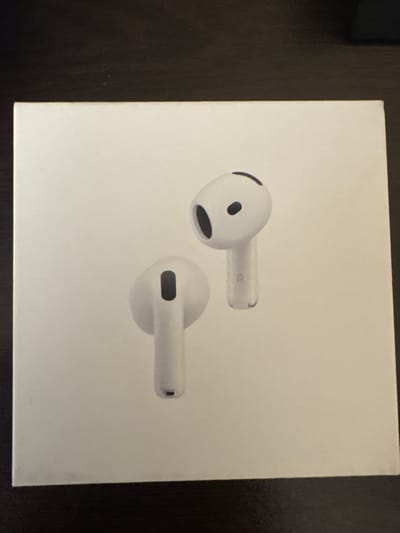 A white rectangular product box, centrally positioned, rests on a dark, subtly textured surface. The box's top surface features a raised, embossed image of two white wireless earbuds, consistent with Apple AirPods 3rd generation. The left earbud, in the foreground, is angled with its stem extending downwards, revealing a dark, oval-shaped speaker opening on its inner side and a dark sensor opening on the stem. The right earbud, positioned slightly behind and to the right, is also angled downwards, with the letter 'R' faintly visible on its translucent stem. A dark oval-shaped speaker opening is also discernible on its inner side. The dark surface, possibly wood or a matte tabletop, extends beyond all edges of the box. The scene's environment lacks specific geographical identifiers, therefore no location information regarding the city of Saedinenie, Bulgaria, or any other specific city, is discernible from the visual content. The lighting is soft and even, resulting in minimal shadows. No human figures or discernible actions are present.