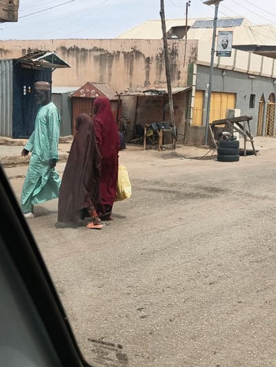 The scene is a daylight street view in Hotoro South, Nigeria, captured from within a vehicle.
In the foreground, a paved asphalt road, light grey with visible aggregate, extends across the frame. Three individuals are walking from left to right. The first, an older male with a white beard, wears a light blue traditional garment and a patterned cap. He is mid-stride, with his left foot in a white slipper visible. Following him is a smaller individual, likely a child, wearing a dark brown full-length garment over patterned leggings and orange sandals. Behind them, an adult female in a dark red full-length garment carries a yellow plastic bag in her right hand; only her feet in dark sandals are visible.
The midground features a collection of commercial structures and buildings. On the left, a dark blue corrugated metal kiosk with a green canopy bears the faded inscription "FORENSIC VENTURE." To its right, a weathered, light brown wall with stains and graffiti ("BA ADAWU") spans the background. Further right, a small brown corrugated metal stall is visible, followed by a wooden makeshift stall or workbench with various items. Behind this stall, faded writing on a wall reads "ZA BATI DIRI MU." To the far right, a grey building with a golden-yellow corrugated metal shutter on the ground floor and a decorative golden-yellow metal gate is visible. The building has a light-colored, possibly tiled, pitched roof.
A wooden utility pole stands on the right, supporting overhead power lines and a metal pole. Attached to the metal pole is a solar panel at the top and a banner below it, displaying "TARAYA TI" and a portrait of a man with additional text (partially visible "DR ALIYU..."). Near the base of the utility pole, three black tires are stacked horizontally, next to which a small internal combustion engine (possibly a generator) rests on a stand. The sky is hazy light blue.