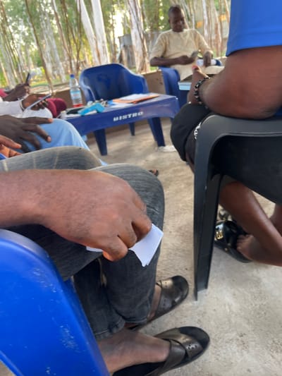 The image shows a group of people seated outdoors, likely in a casual setting. The subjects are primarily visible from the waist down, with some hands and arms also in frame. One person in the foreground is holding a small piece of white paper. Another individual is wearing a light-colored traditional garment and appears to be holding a phone. Several people are wearing dark-colored sandals or flip-flops.
The seating appears to be on blue plastic chairs, some of which have the word "BISTRO" printed vertically in white text on them. A partially visible bottle of water is also on one of the chairs. The ground is a light-colored concrete or paved surface. In the background, there are thin, vertical structures that resemble trees or reeds, suggesting an outdoor, possibly natural or semi-natural environment.
The lighting suggests it is daytime, with bright sunlight filtering through the trees in the background. No specific emotions are discernible from the cropped view. The overall impression is one of a relaxed gathering or meeting. The location context provided is Mayo Dasa, Nigeria, which fits the visual cues of an outdoor setting.