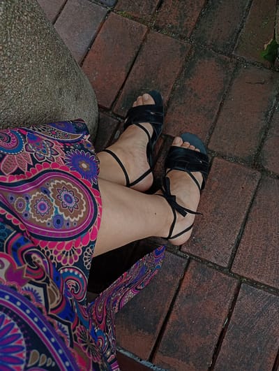 The image shows a close-up, overhead view of a person's legs and feet, wearing black lace-up sandals. The person is wearing a colorful dress with a paisley pattern in shades of pink, purple, blue, and brown. The legs are resting on a brick patio, which is reddish-brown in color and appears weathered. To the left of the legs is a large, rough, textured, greyish-brown stone, possibly a boulder or part of a bench. The overall scene suggests an outdoor setting, possibly a garden or a public space in Constance, Germany, where such paving and seating arrangements are common. There are no visible identifying features of the person.