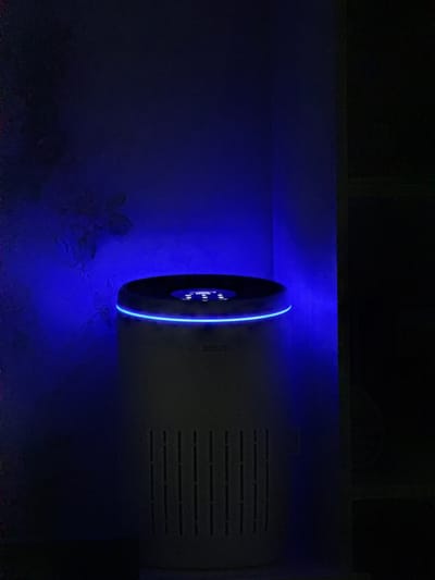 The image shows a white Bosch air purifier with a blue LED light ring around its top. The device is placed in a dark room, likely a residential interior, with a blue light illuminating the wall behind it. The wall appears to have a subtle floral pattern. The air purifier has a slatted vent on its lower front. The scene is not identifiable as being located in Slavyani, Bulgaria or any other specific city. There are no people or interactions depicted.