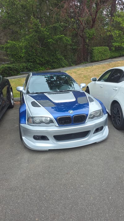 This is a close-up, eye-level, outdoor, vertical shot of the front of a custom BMW M3. The car is silver and blue with a wide body kit and aftermarket hood vents. It is parked on asphalt, with grass and trees visible in the background. A white car is parked to the right of the BMW, and a black car is partially visible to the left. The scene is an overcast day. The exact location in Lynnwood, Washington, USA is not determinable from the image alone.