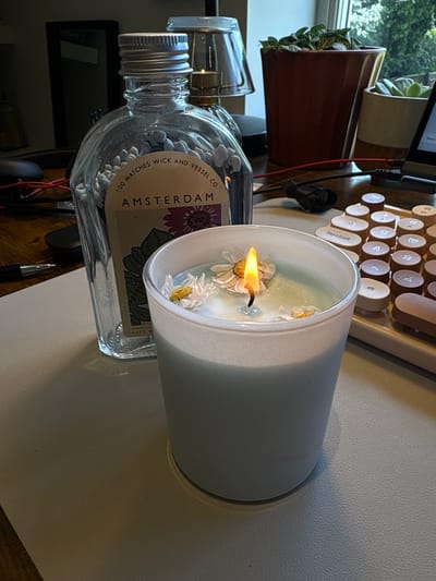 A lit candle with daisies floating in the wax sits on a desk next to a bottle of matches. The candle is in a white cylindrical container. The match bottle is clear glass with a silver lid and a label that reads "100 MATCHES WICK AND VESSEL CO. AMSTERDAM". In the background are plants in terracotta pots, a vintage-style keyboard, and what appears to be a desk or table surface. The scene is indoors. The location in the city of Ipswich, United Kingdom is unknown.