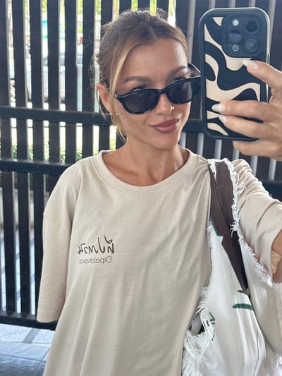 The image shows a woman taking a selfie in Tong Yang, Thailand. She is wearing sunglasses and a light-colored t-shirt with a graphic design and text on it. The woman is standing in front of a dark, slatted fence. She is holding up a smartphone with a patterned case in her right hand. A tote bag with a similar design is visible on her left side. The lighting suggests it is daytime, and the background appears to be outdoors.