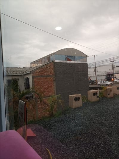 The scene captures a view through a window on an overcast day. Outside, there is a building complex with a gray wall structure and a warehouse-like building with an arched roof, bearing the word "XAMIR" on it. A smaller building with a sloped roof is also visible. In the foreground, there are concrete blocks with tire inserts. A SOAT sign and gravel cover the ground. Cars are seen in the distance on a road.