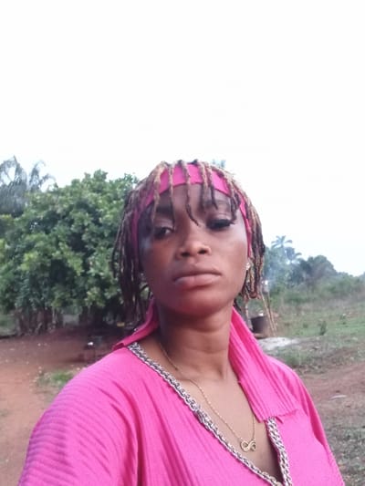 A young woman is depicted in a portrait-style photograph, captured outdoors in what appears to be Ogbe, Nigeria. She is positioned on the left side of the frame, gazing directly at the camera with a neutral expression.
She is wearing a bright pink garment, possibly a top or dress, featuring a textured, pleated fabric and a decorative silver-toned trim along the neckline. A delicate gold chain necklace with a small, gold pendant, resembling a stylized letter 'S' or an infinity symbol, hangs around her neck. Her hair, styled in dreadlocks or braids, is partially covered by a pink band or scarf worn across her forehead.
The background features lush green trees and dense foliage, suggesting a natural or semi-natural environment. The ground closer to the subject appears to be unpaved earth. A significant portion of the right side of the background is dominated by a bright, almost white sky, indicating it is daytime with strong sunlight or overcast conditions. The overall activity appears to be posing for a casual outdoor photograph. There is no visible text present in the image.