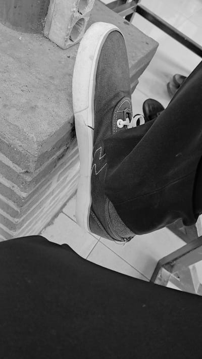 In Julikeude Dua, Indonesia, a person's foot, clad in a dark canvas sneaker with white laces and a white sole, rests on a rough, textured brick structure. The shoe has a distinctive white zig-zag pattern on its side. The person is wearing dark trousers. The background includes a tiled floor and hints of other footwear, suggesting an indoor or covered outdoor setting. The image is in black and white, focusing on the shoe and its immediate surroundings.