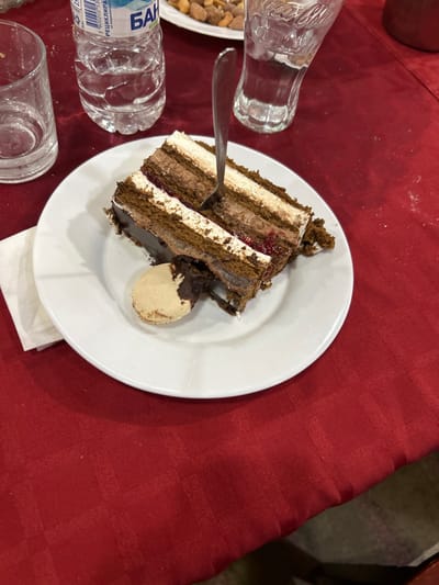 The image depicts an indoor dining scene focused on a dessert. A slice of multi-layered chocolate cake, featuring brown cake layers, white and light brown cream fillings, and a thin red fruit preserve layer, rests on a white circular plate. A silver fork is inserted vertically into the cake. Adjacent to the cake on the plate is a light-colored macaron with a partially visible chocolate filling. A crumpled white paper napkin lies to the left of the plate. In the background, a partially filled clear plastic bottle of "БАН" branded water is visible on the upper left, next to an empty clear glass tumbler. To the upper right, an empty clear glass with an embossed Coca-Cola logo is present. A white plate containing assorted nuts is partially visible in the far upper background. The items are arranged on a red tablecloth with a subtle woven checkered pattern. No individuals are visible. The specific location within Arkovna, Bulgaria, cannot be determined from the visual information provided.